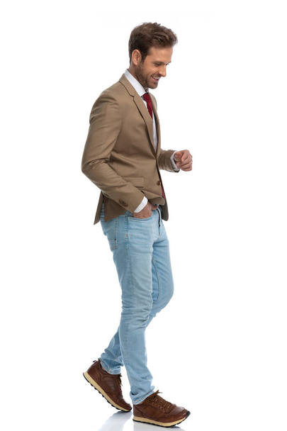 side view of a sexy businessman walking, looking down, smiling and holding one hand in pocket  - Fotó, kép