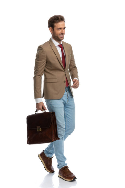 side view of a handsome businessman walking on his way with a briefcase and a smile on his face - Fotó, kép