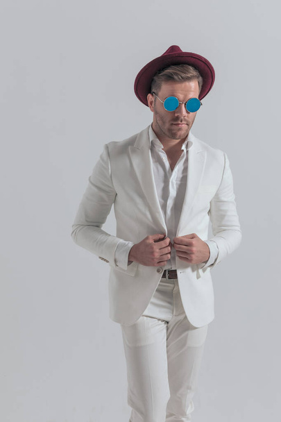 cool fashion model fixing his jacket, wearing a hat and sunglasses against gray background - Fotó, kép