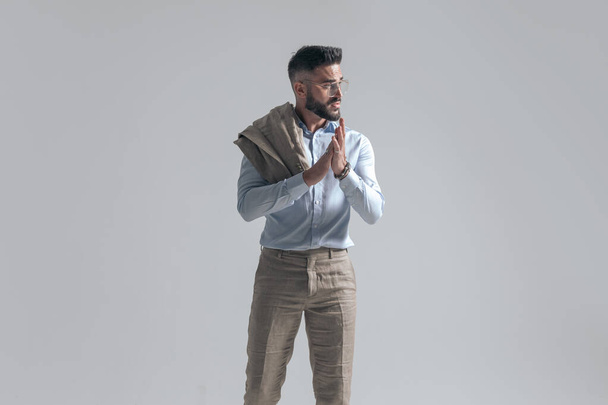 retrato de atractivo joven hombre de negocios con chaqueta sobre el hombro frotando las palmas y mirando a un lado mientras está de pie sobre fondo gris en el estudio - Foto, Imagen