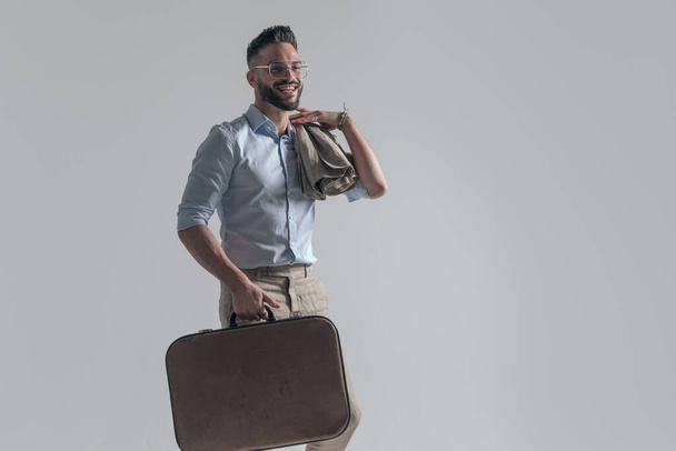 happy young man with jacket over shoulder holding suitcase and smiling while looking to side on grey background in studio - 写真・画像