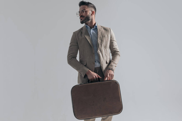 cool bearded young model holding briefcase and looking to side while walking on grey background and posing in studio - Foto, afbeelding