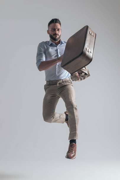 cool young fashion model leaping up while holding jacket and briefcase on grey background in studio - Foto, Bild