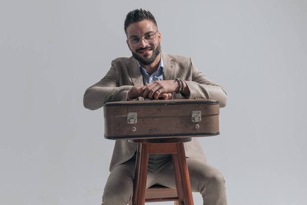 happy young businessman smiling and holding arms on suitcase while sitting on grey background in studio - Фото, изображение