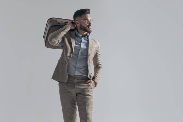 cool businessman with glasses holding hand in pocket and bag over shoulder while looking to side on grey background in studio - Foto, afbeelding
