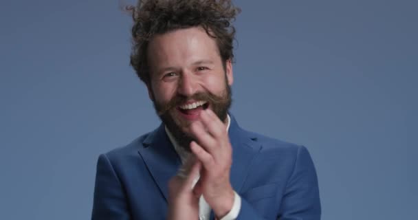excited businessman with curly hair in studio clapping, being happy, smiling, pointing fingers and laughing on blue background - Záběry, video