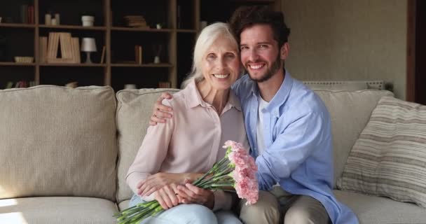 Older woman holds flowers sit on sofa with grownup son - 映像、動画