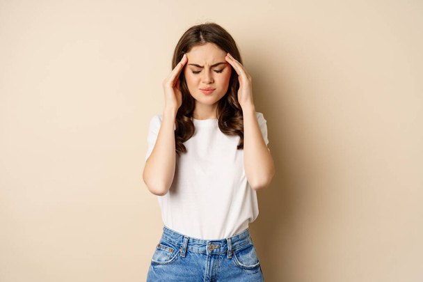 Health and women concept. Portrait of girl feeling sick, touching head temples, feeling headache, migraine, standing over beige background - Photo, Image