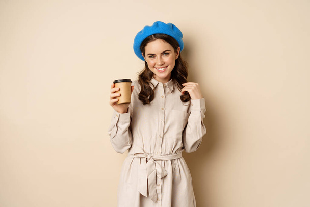 Hermosa chica moderna posando con una taza de café para llevar, bebiendo de la taza reutilizable de la cafetería y posando feliz, fondo beige estudio - Foto, Imagen
