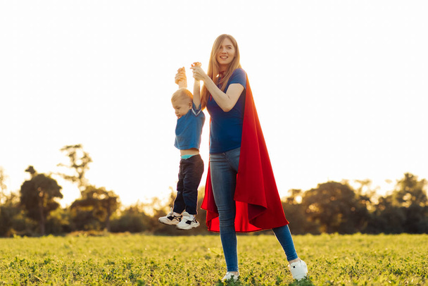 Mother and her child play together outdoors. Mom and son in superhero costumes. Mom and baby have fun, smile and hug. Family vacation and togetherness. Concept of Mother's Day, family - Fotoğraf, Görsel