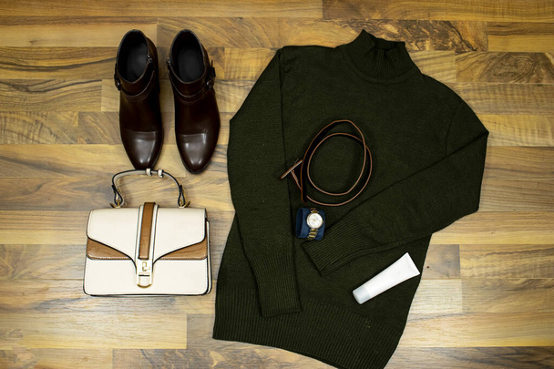 Un conjunto de ropa y accesorios de moda. suéter verde y tubo cara crema, bolso de mano, botas de tobillo en la mesa de madera, disposición plana. Estar en tendencia - Foto, imagen