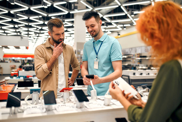 Sales consultant advises man who chooses new smartphone in store of household appliances, electronics and gadgets. Professional consultation in store. Sale Day. - Φωτογραφία, εικόνα