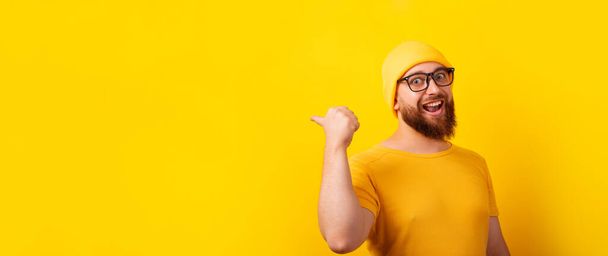 sonriente hombre mostrando gesto pulgar hacia arriba y señalando el espacio vacío sobre el fondo amarillo, diseño panorámico - Foto, imagen