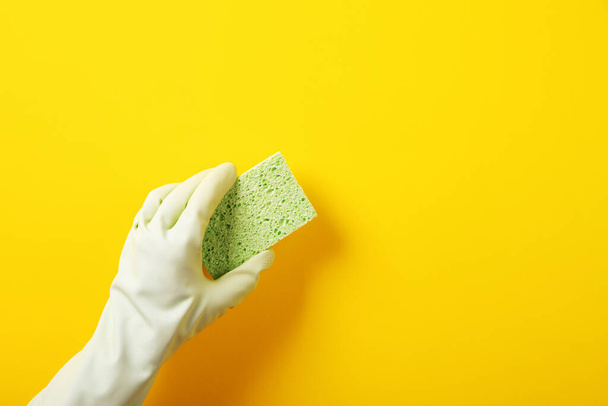 A hand in a green rubber glove holds a washing sponge over yellow background - Фото, изображение