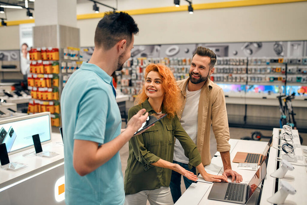 Young couple receives advice from seller in store of household appliances and gadgets, buying new laptop. Male consultant helps in choosing laptop. - Foto, Imagen