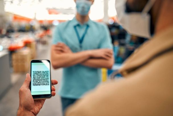 Man and seller in protective masks holds smartphone, on screen of which QR code in store of household appliances, electronics and gadgets. Payment by QR-code, online stores, cashless technology. - Fotó, kép