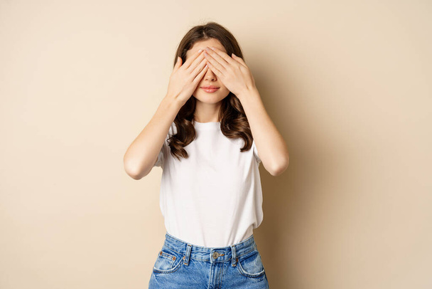 Surprise. Happy young woman shut eyes with hands and waiting for gift, standing over beige background - Photo, Image