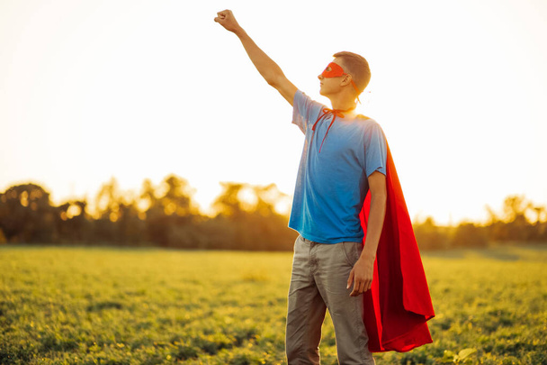 Joyful courageous man in a superhero costume poses on the field against the backdrop of sunset. A man in a red cape of a superhero makes a victory gesture, a man in the image of a superhero grows up - Foto, Imagen