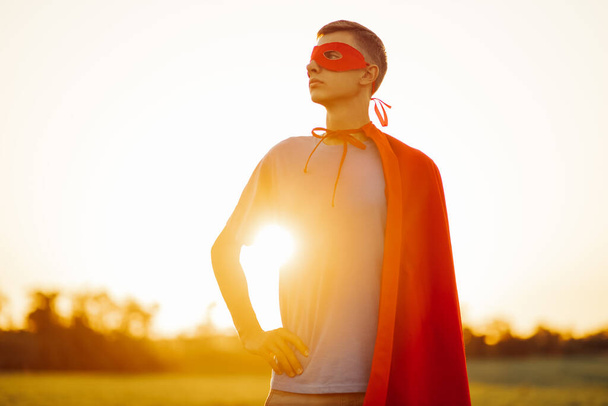 Happy man in a red cape of a superhero and a mask on his face, Strong confident man against the backdrop of a sunset sky in nature. concept of power, strength and people - Zdjęcie, obraz