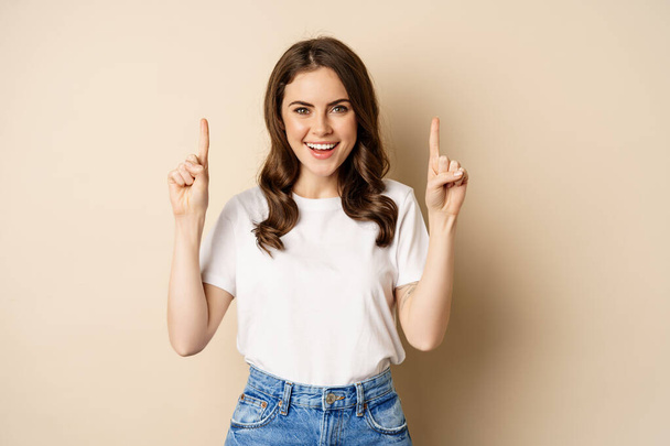Mujer joven entusiasta, cliente femenino señalando con los dedos hacia arriba y sonriendo, mostrando pancarta o logotipo, de pie sobre fondo beige - Foto, Imagen