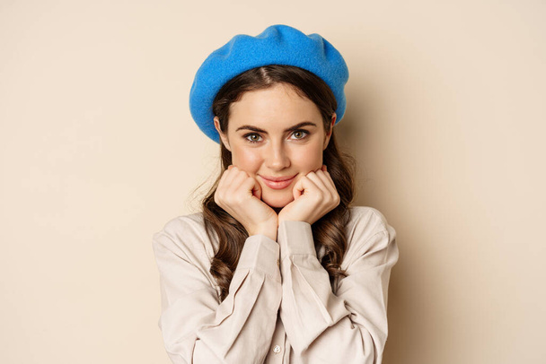 Close up portrait of feminine young woman in trendy french hat, posing cute and romantic, making silly coquettish face and tender smile, beige background - Fotó, kép