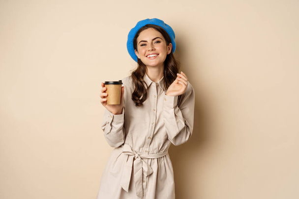 Beautiful happy girl drinking takeaway coffee from cafe and smiling, posing with cup of beverage, beige background - 写真・画像