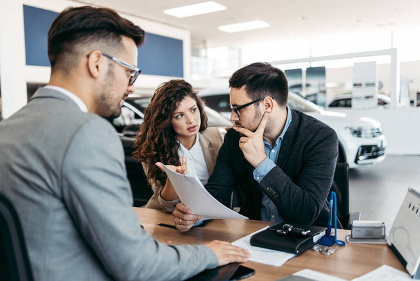 Middle age couple choosing and buying car at car showroom. Car salesman helps them to make right decision. Man signs buyers contract. - Φωτογραφία, εικόνα