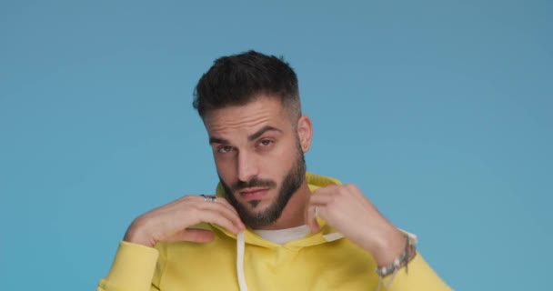 young casual man putting his hood on and smiling at the camera on blue background - Záběry, video