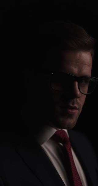 project video of handsome unshaved businessman with glasses looking to side and grimacing while standing in the dark in a mysterious background studio - Felvétel, videó