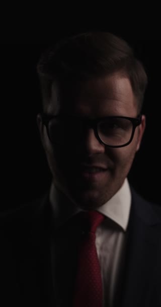 happy young businessman in suit wearing glasses, fixing tie and smiling on black background in a mysterious studio - Felvétel, videó