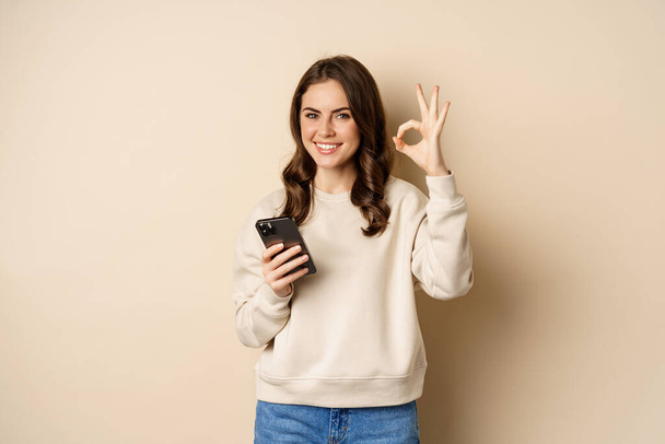 Красива жінка тримає мобільний телефон, мобільний телефон і знак, рекомендує додаток, додаток для покупок, стоїть над бежевим тлом
 - Фото, зображення