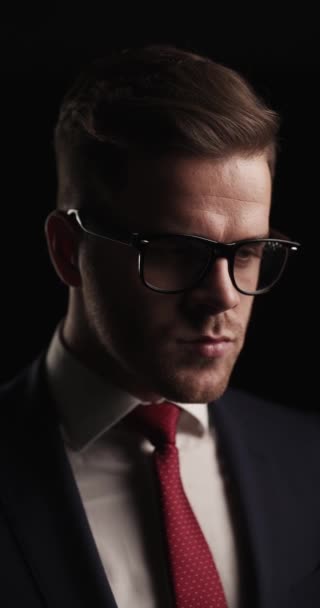 vista lateral del hombre sexy con gafas mirando hacia otro lado, siendo considerado y pensando mientras está de pie sobre fondo negro en el estudio - Metraje, vídeo