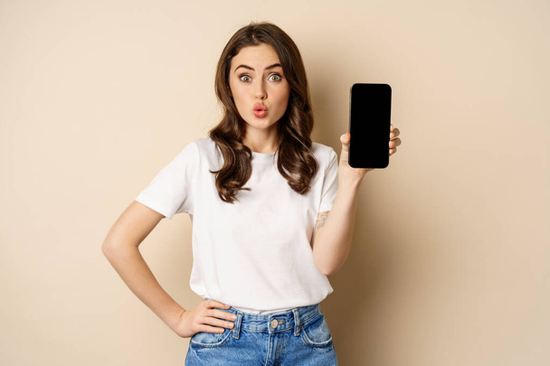 Online shopping and app concept. Young woman smiling and showing mobile phone screen, application interface, standing over beige background - Фото, изображение