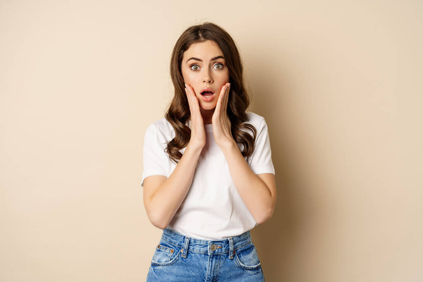 Portrait of stylish young woman gasping, looking surprised and amazed, impressed by smth, standing in t-shirt and jeans over beige background - Photo, image