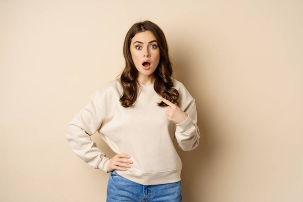 Surprised girl pointing at herself and look with disbelief, standing over beige background - Фото, изображение