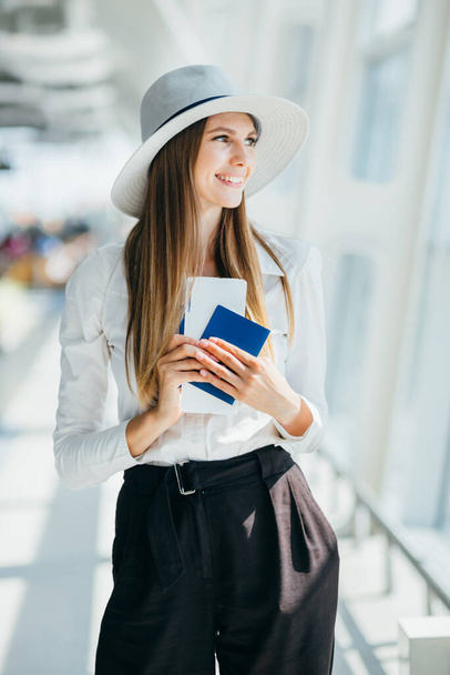 Una alegre mujer de negocios esperando su vuelo en el aeropuerto. Una joven en el aeropuerto. Retrato de una joven feliz con pasaporte y tarjeta de embarque en el aeropuerto - Foto, Imagen