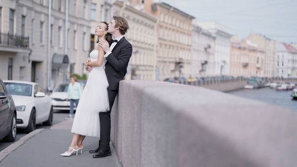 Side view of caring lovely just married couple embracing by the granite sidewalk. Action. Elegant bride and groom outdoors in the street. - Фото, изображение