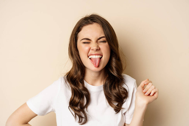 Close up of happy, carefree beautiful woman posing silly, showing tongue, having fun, standing in casual white t-shirt against beige background - Photo, image