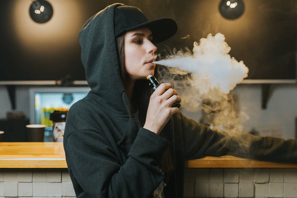 Egy fiatal csinos nő sapkában elszív egy elektromos cigarettát a gőzboltban. Hip-hop stílusban. Közelkép. - Fotó, kép