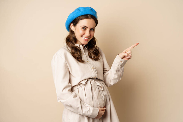 Young future mother, pregnant woman smiling, posing with big belly third trimester of pregnancy, looking happy, expecting baby, pointing right at logo, banner or advertisement - Photo, Image
