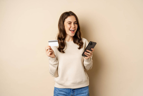 Menina sorrindo usando aplicativo móvel, compras de smartphones e cartão de crédito, de pé sobre fundo bege, ordem smth - Foto, Imagem