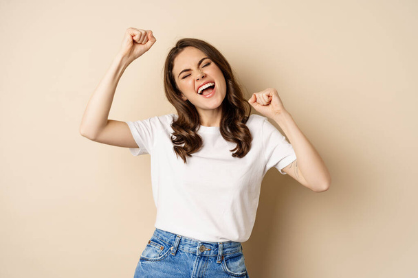 Enthusiastic adn surprised woman winning, dancing and celebrating, standing in tshirt over beige background - Фото, изображение