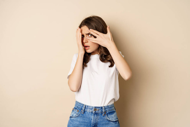 Young woman scared to watch, cover eyes with hands and peeking through fingers at scary moment, standing over beige background - Foto, Imagem