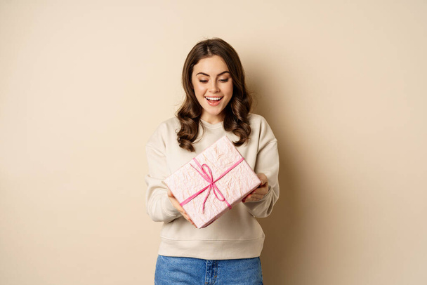 Emocionado hermosa chica con caja de regalo envuelta en rosa, recibir regalos, de pie sobre fondo beige - Foto, imagen