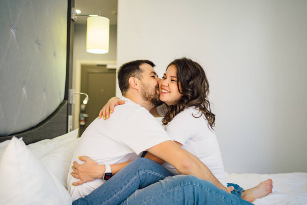 a pregnant woman and a man in white T-shirts and jeans on bed. - Fotografie, Obrázek