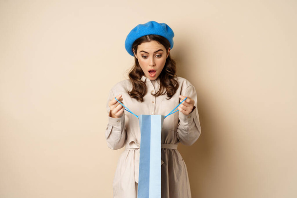 Image of girl looking surprised after opening gift bag, looking at present amazed, standing in stylish outfit over beige background - Фото, изображение