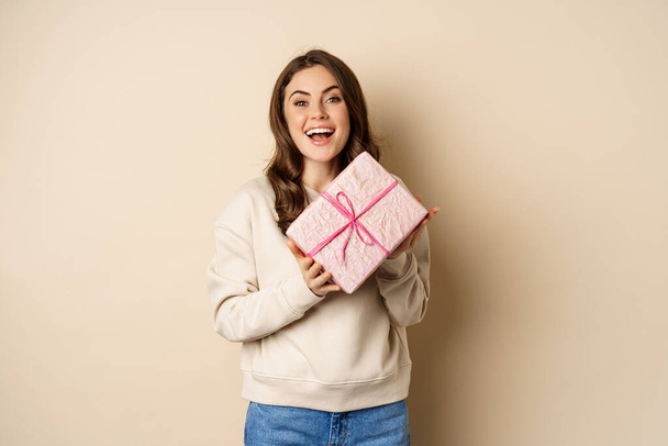 Happy beautiful girlfriend holding pink gift box and laughing, smiling joyful, concept of holidays and celebration, beige background - Photo, image