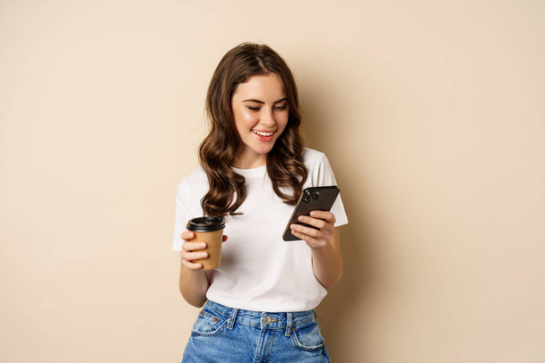 Smiling woman using mobile phone app and credit card, concept of online shopping, contactless payment and delivery, standing over beige background - Foto, afbeelding