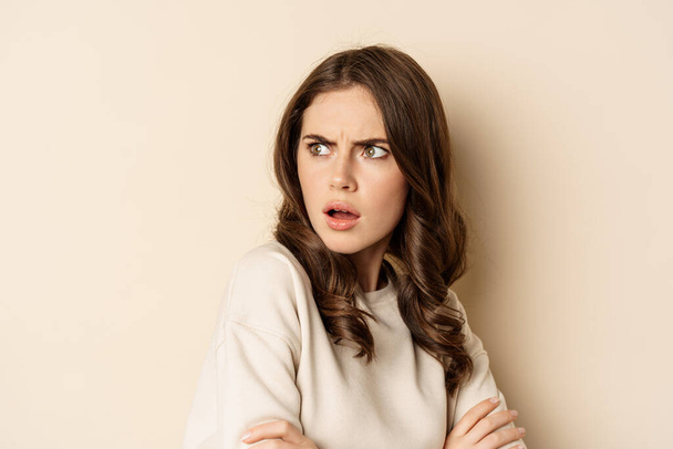 Close up of confused young woman, looking behind with skeptical shocked face expression, standing against beige background - Фото, изображение