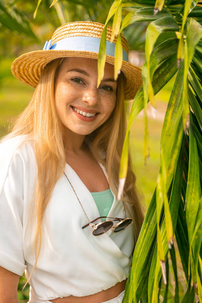 Close-up portrait of a beautiful amazing luxurious blonde woman with a straw hat standing under a palm tree on the beach, relaxing on a tropical island - 写真・画像